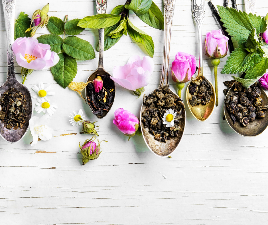 Silver spoons holding tea leaves on a rustic table with flowers and mint leaves sprinkled throughout.